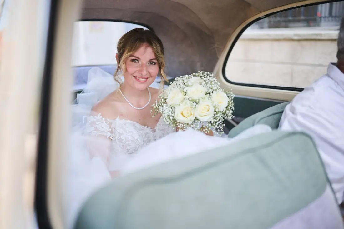 La mariée souriante à l'arrière d'une voiture ancienne, bouquet de roses blanches